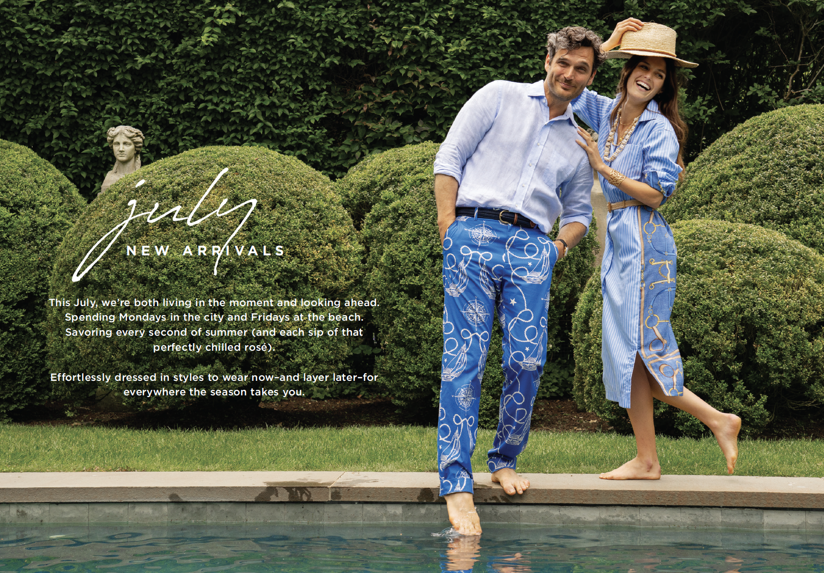 A man and woman in blue summer clothing standing by a swimming pool, smiling, with manicured bushes and a garden statue in the background.