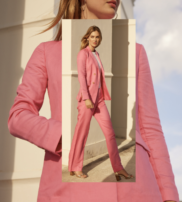 A woman standing outdoors in a pink pantsuit with a blazer and matching trousers, posing against a beige wall with sunlight and clouds visible in the background.