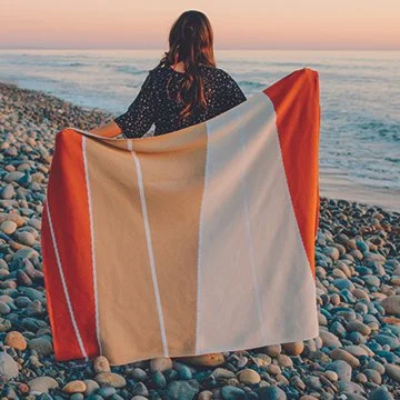A woman sitting on a pebble beach holding a large striped towel, facing the ocean at sunset.