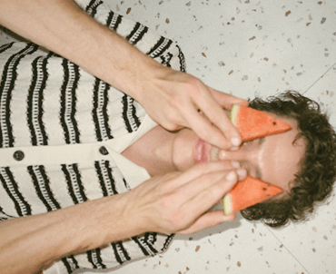 Person holding slices of watermelon over their eyes while lying on the floor.