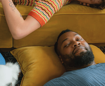A man lying on a yellow pillow with his eyes closed, and a woman leaning over him on a yellow couch, wearing a colorful striped shirt. Part of a white and black pet is visible at the bottom left corner.