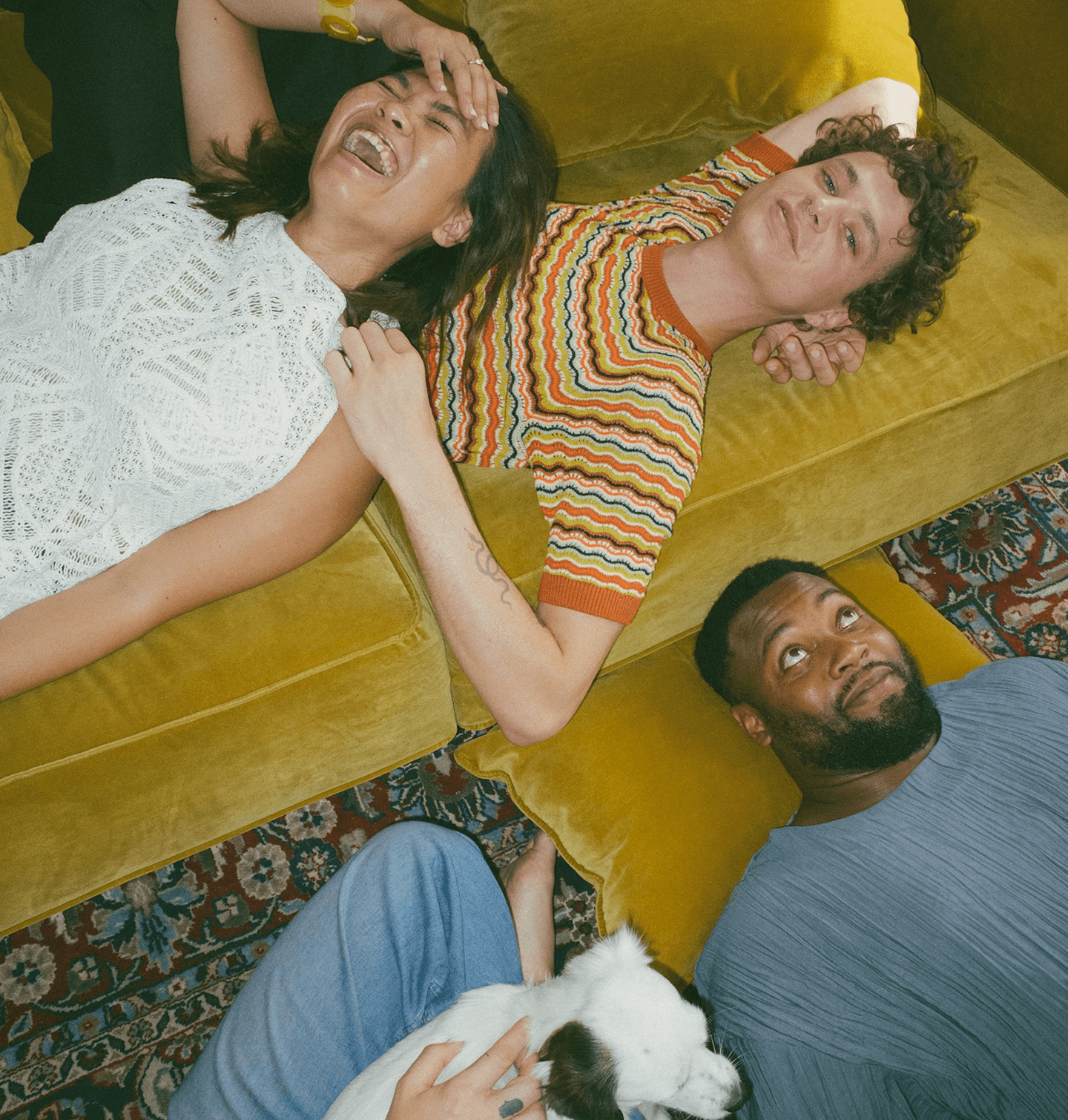 Three people lying on a yellow couch and floor, smiling and laughing, with a white dog on someone's lap, in a cozy, colorful living room.