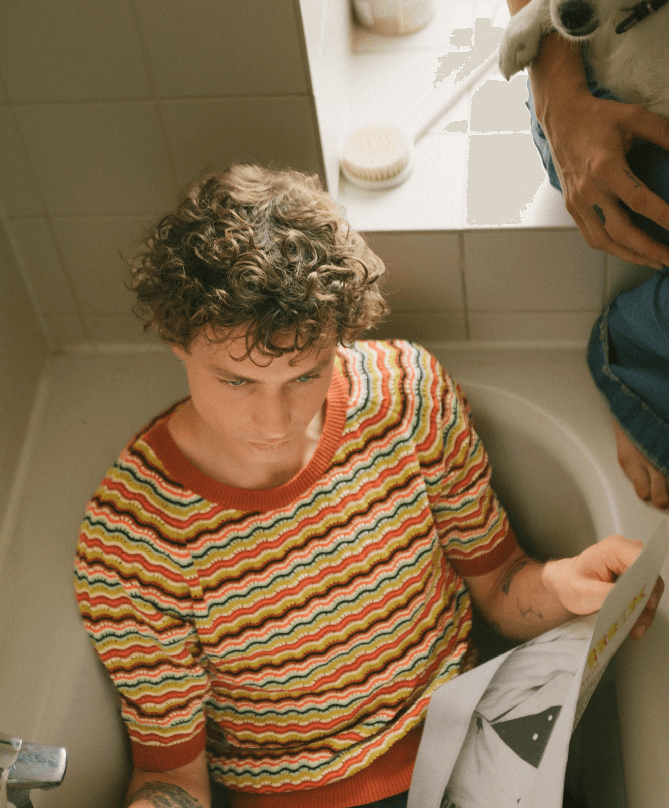 A person with curly hair sitting on a bathroom toilet, looking down at a piece of paper. Another person is holding a small dog nearby.