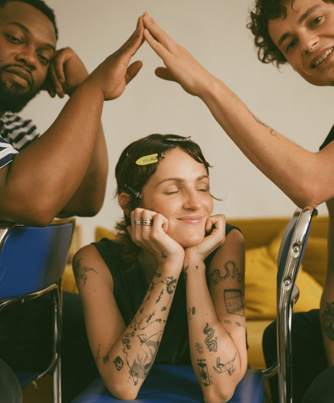 Three friends smiling, with two making a high five over a person with tattoos sitting with eyes closed and a content smile.