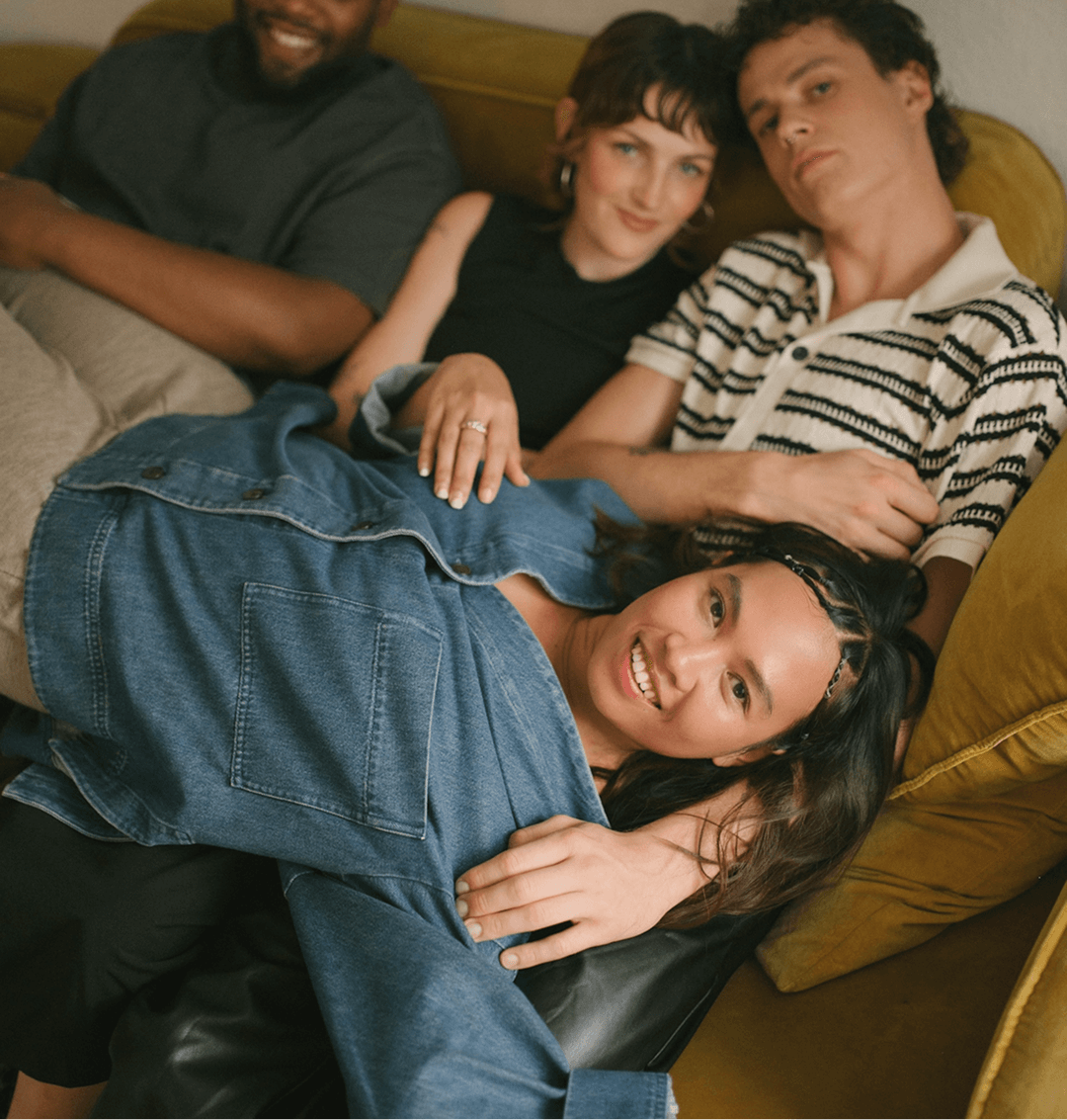 Four young adults relaxing together on a beige couch, smiling and looking at the camera, with one lying down and three sitting close.