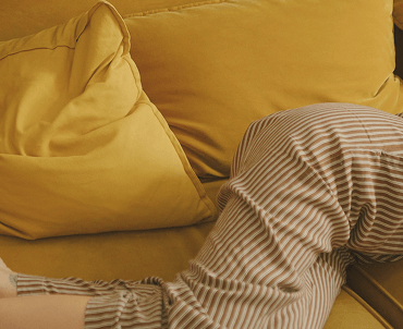 Close-up of a person's legs wearing striped pajama pants lying on a yellow couch with yellow pillows.