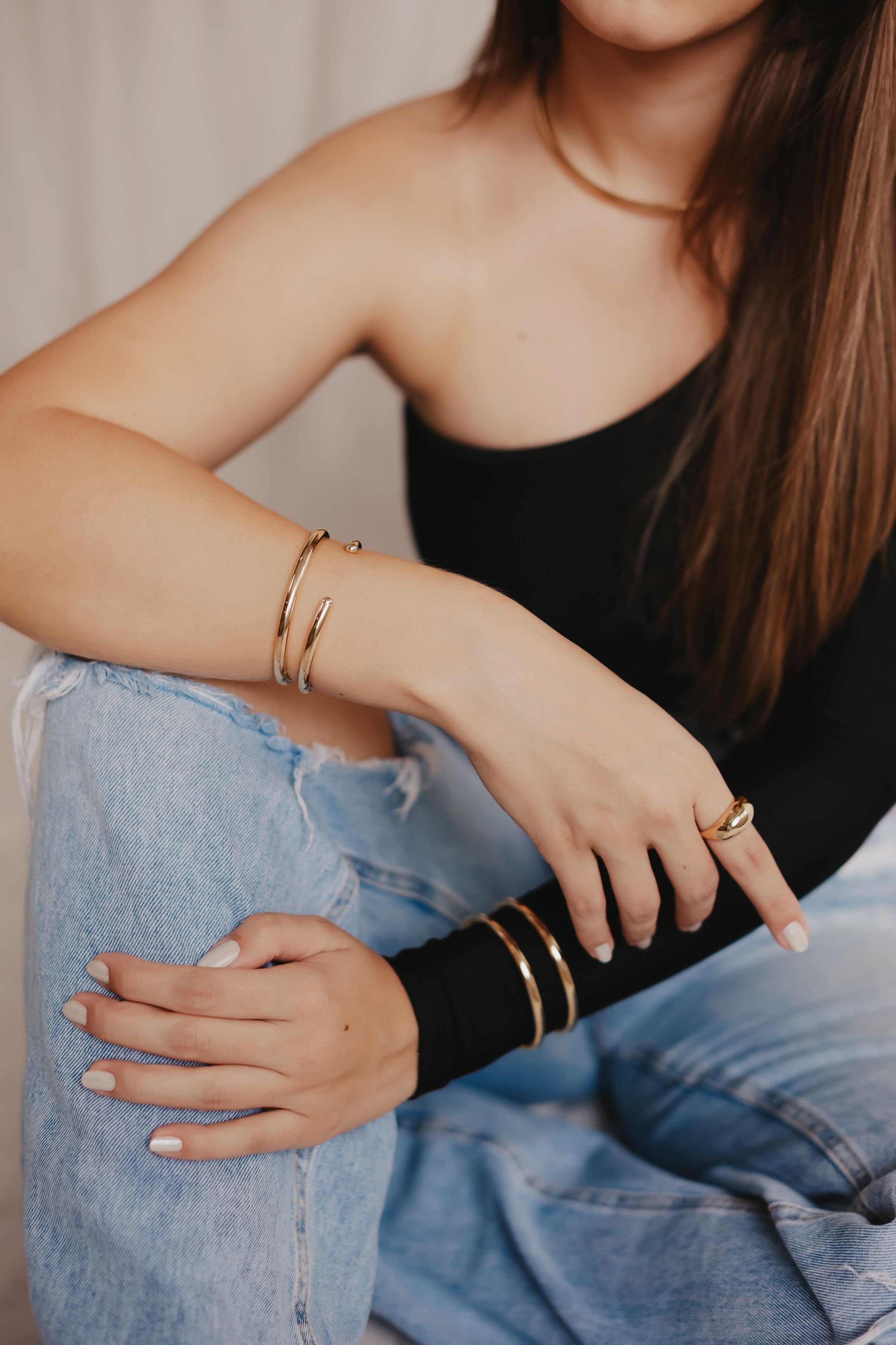 Close-up of two people, one in a black top with long straight hair, arms crossed, wearing gold and black bangles, and rings, and the other in distressed denim jeans with hand visible, wearing rings and a bracelet, posing together.
