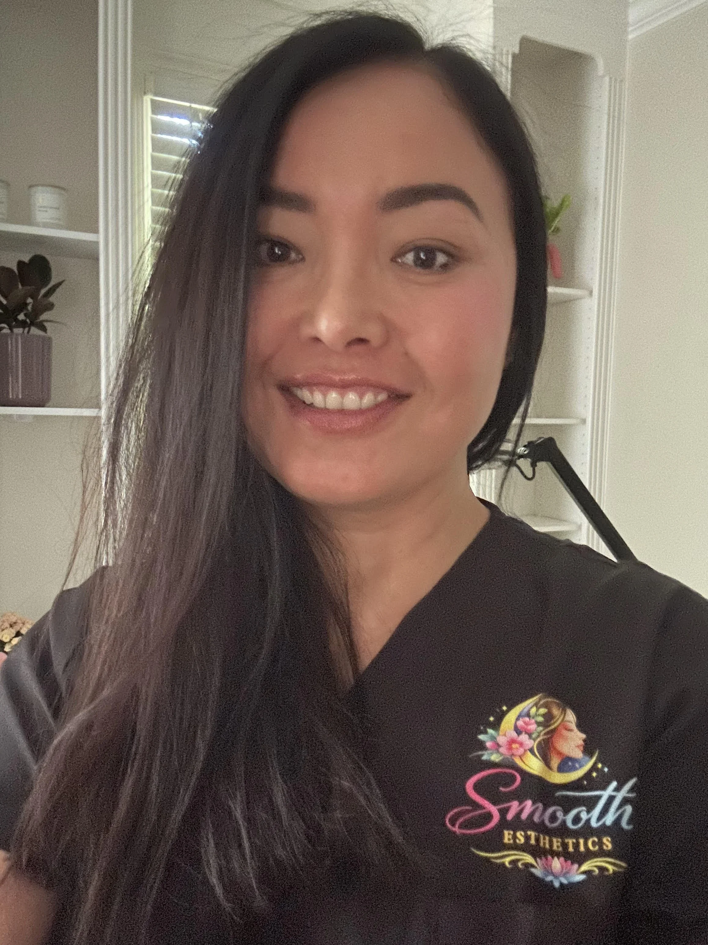Close-up of a woman with long, dark hair, smiling, wearing a black shirt with a colorful logo for an esthetics business that features a woman with flowers in her hair.