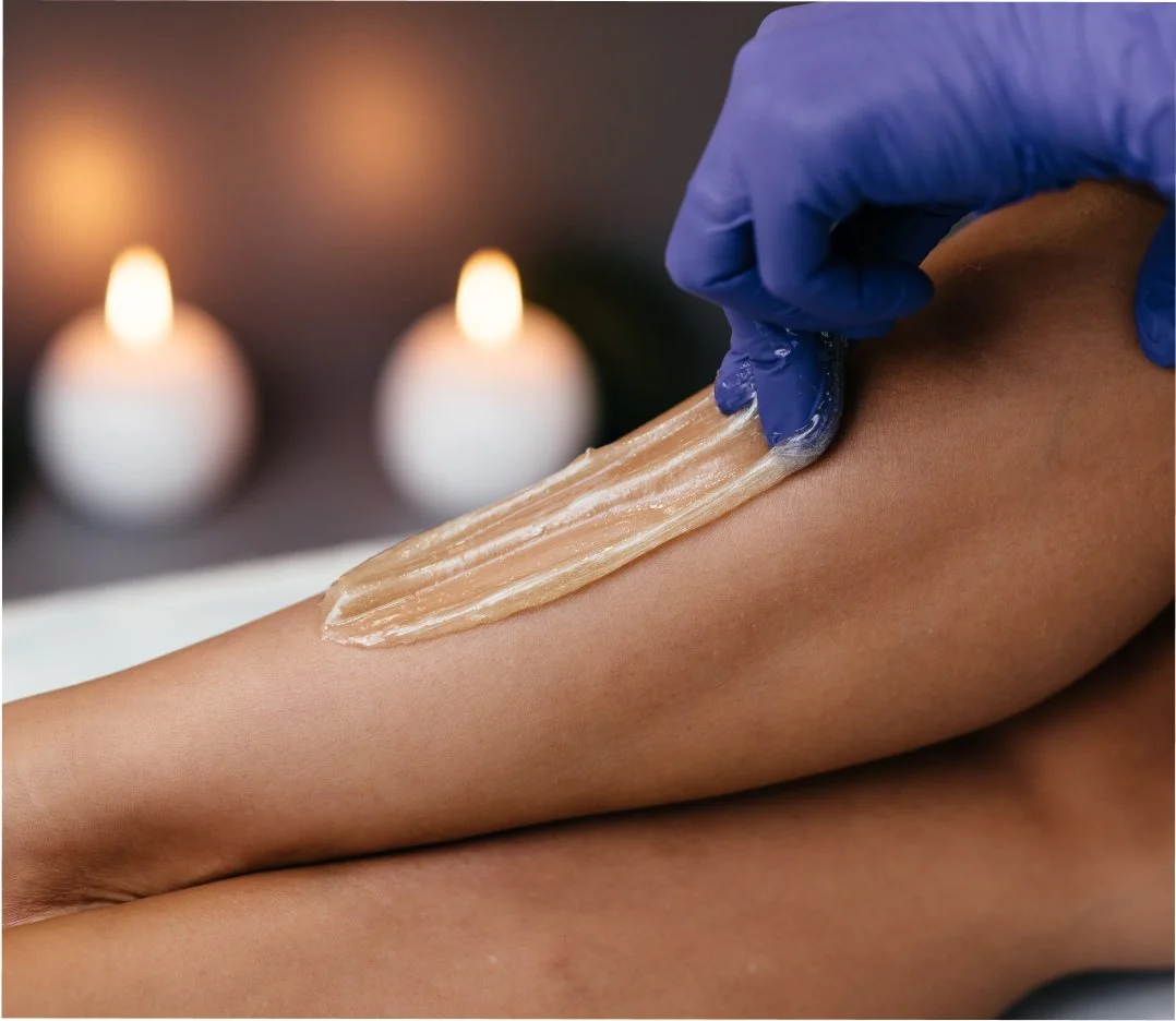 A person is receiving a waxing treatment on their leg, with wax being applied by a professional wearing blue gloves. In the background, there are two lit candles creating a relaxing atmosphere.