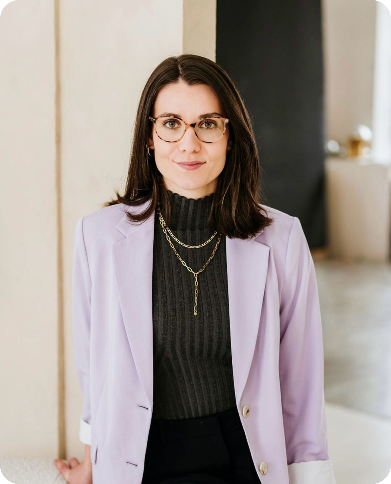 Clara Crombach mit dunklen Haaren, Brille, schwarzem Rollkragenpullover, lila Blazer und Halsketten, vor einem neutralen Hintergrund.