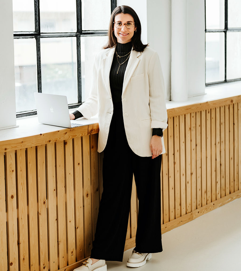 Clara Crombach steht in einem hellen Raum am Fenster, neben ihr ein Laptop auf der Fensterbank – sie trägt eine Brille und ein Business-Outfit mit einem hellen Blazer.