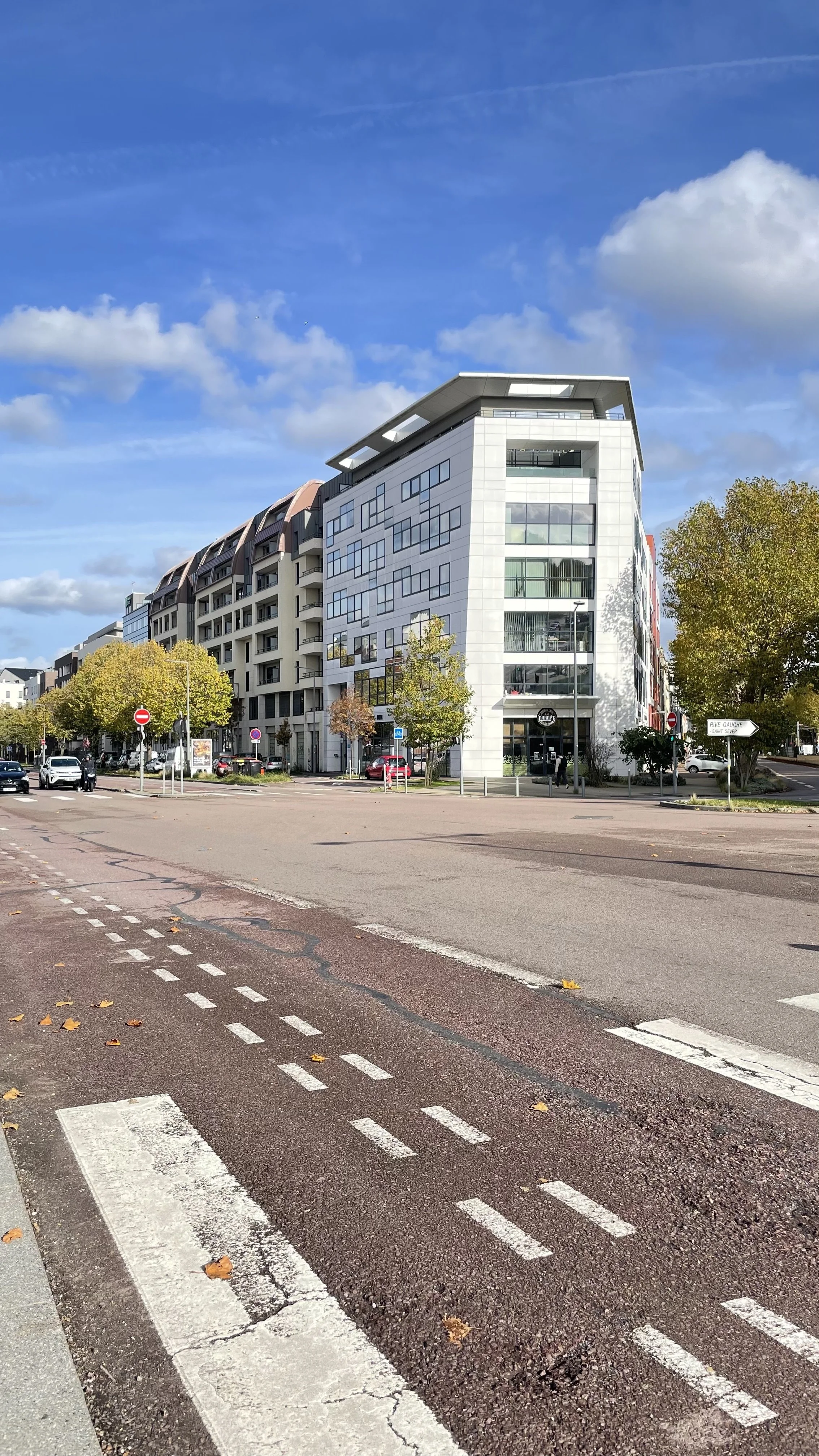 Photo d'une rue urbaine avec un bâtiment moderne blanc et en verre, des voitures stationnées, des arbres avec des feuilles jaunes, et un ciel bleu avec quelques nuages.