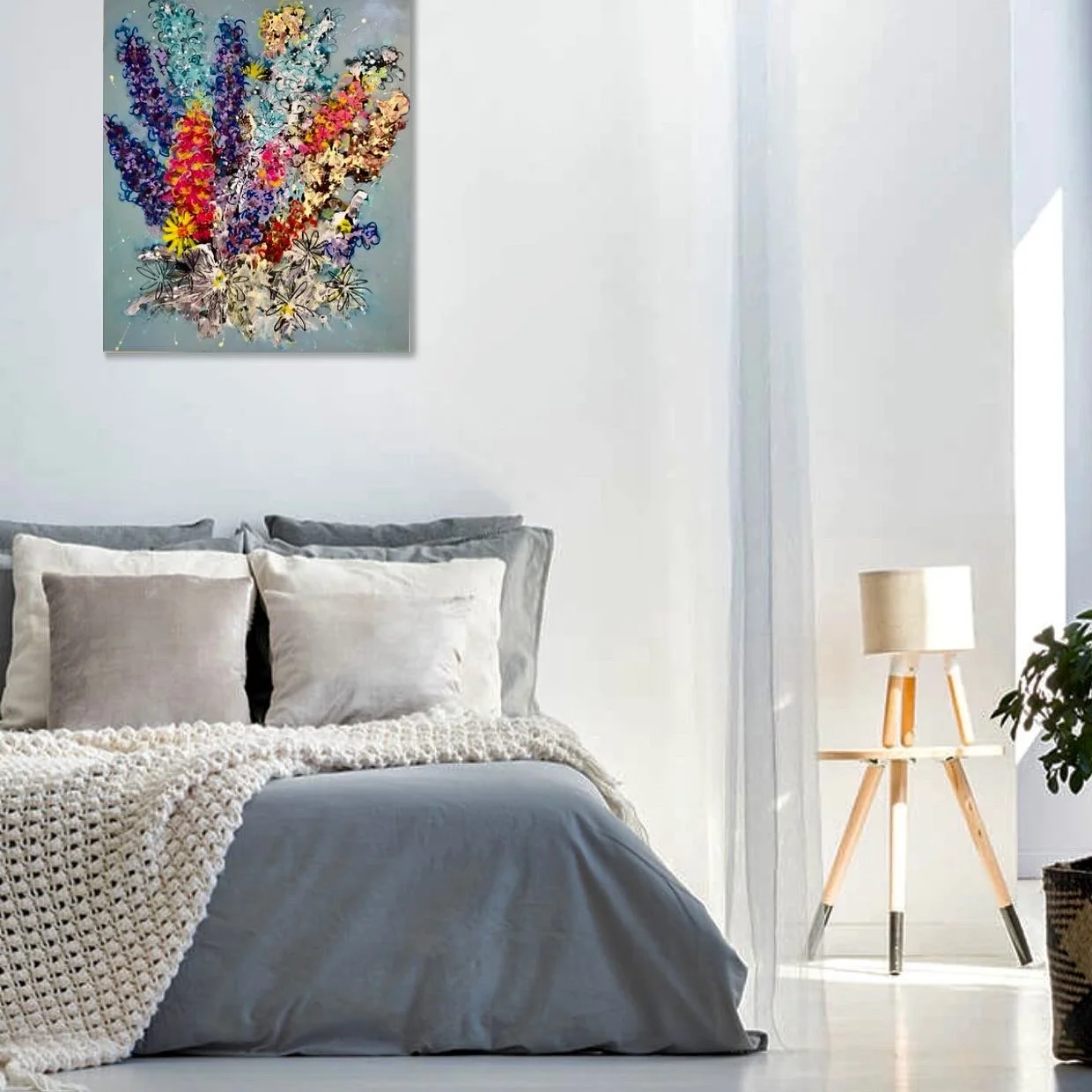 A bedroom with a neatly made bed with gray and white pillows, a cream-colored crocheted blanket, and a gray bedspread. There is a wooden tripod floor lamp with a cream lampshade beside the bed. A colorful abstract floral artwork hangs on the white wa