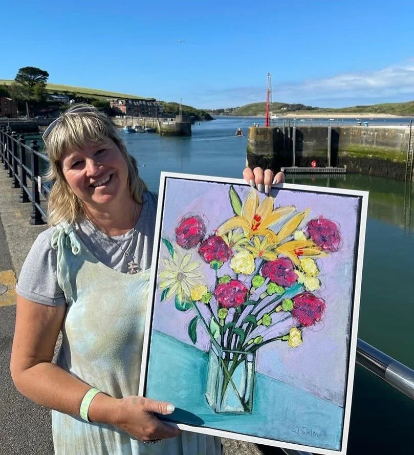 Woman smiling and holding a colorful painting of a flower bouquet in a glass vase, outdoors near water with a pier and hills in the background.