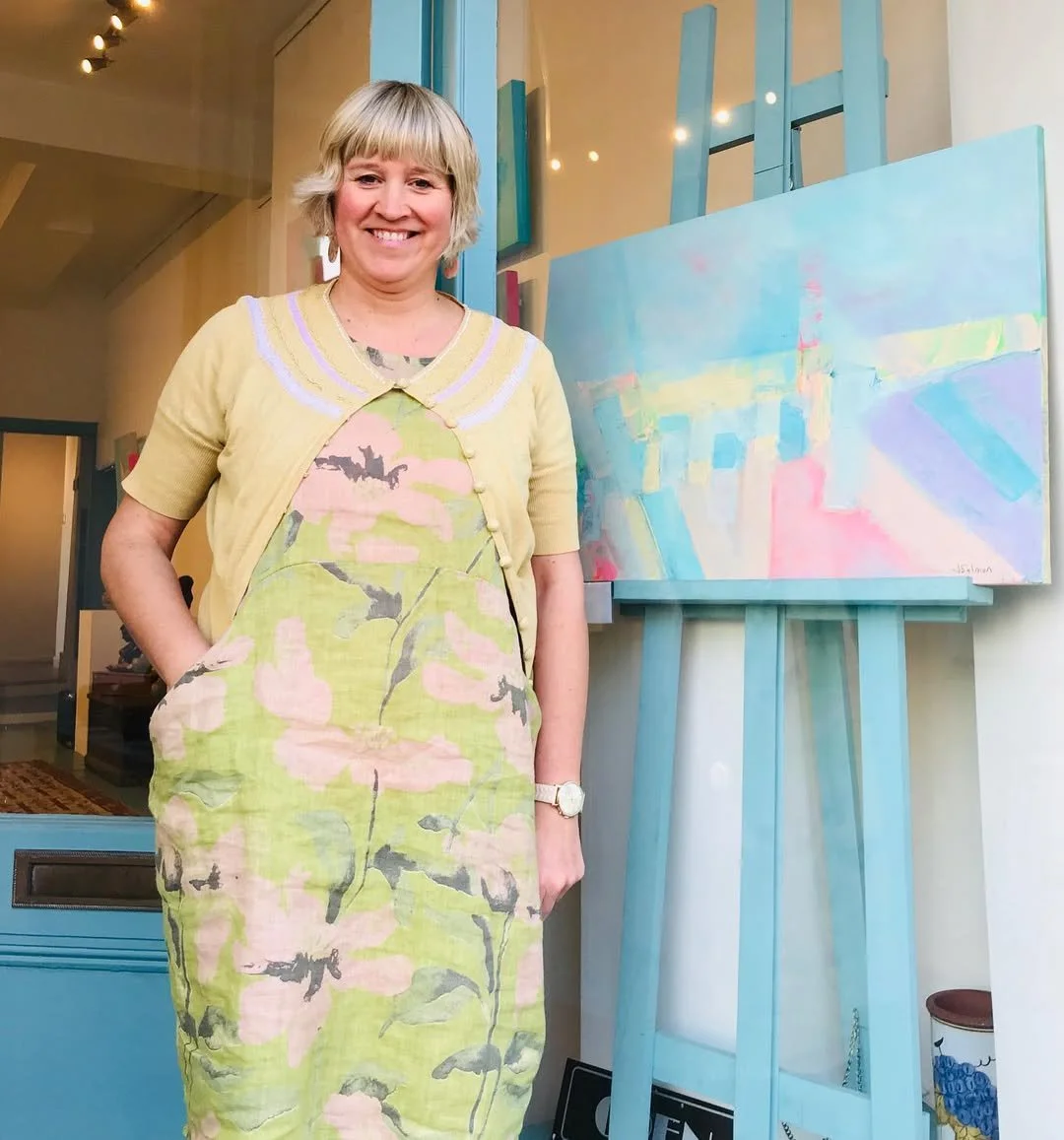 A smiling woman standing next to an abstract pastel-colored painting on an easel in an art gallery.