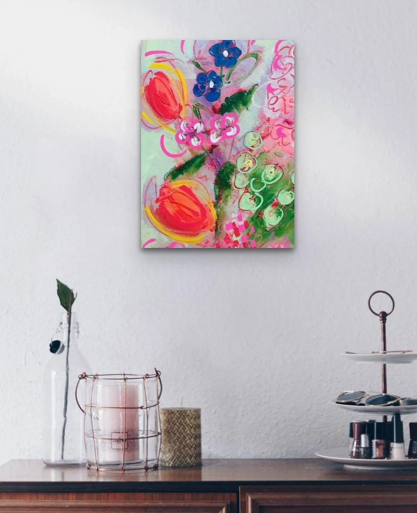 Colorful abstract floral painting hanging on a white wall above a wooden table with various decorative items including a tall vase with a single leaf, a candle in a wire holder, and a two-tiered tray with accessories.