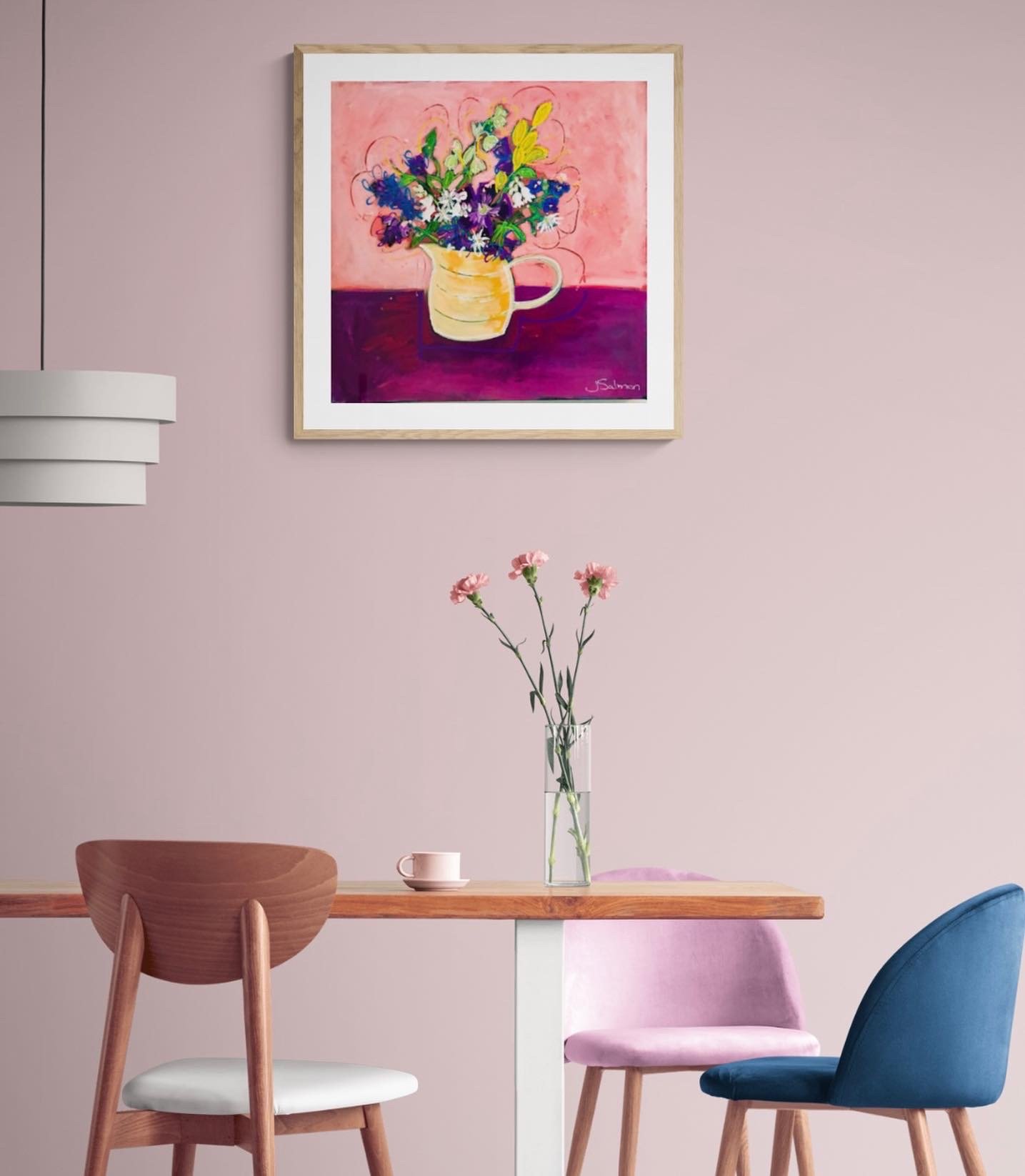 Interior of a room with a light pink wall, a wooden dining table, and three chairs—one brown, one pink, and one blue. On the table, there is a clear glass vase with pink flowers and a small pink cup. Above the table, a colorful painting of a bouquet 