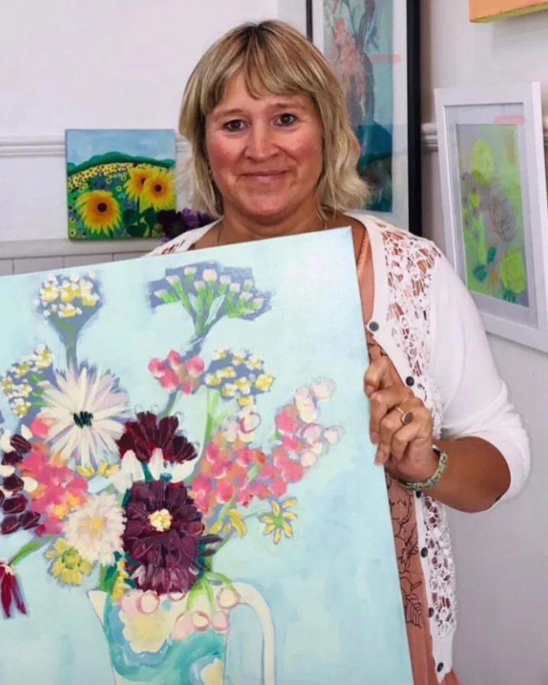Woman with blond hair holding a colorful floral painting in an art gallery or studio, with other paintings of sunflowers and abstract art on the walls.