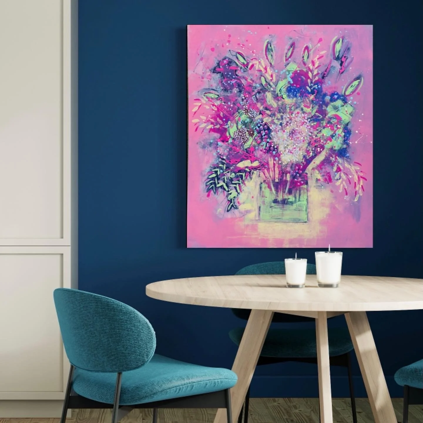 A modern dining area with a light wood round table, teal velvet chairs, and two white candles. A vibrant pink and purple floral painting hangs on a dark blue wall.