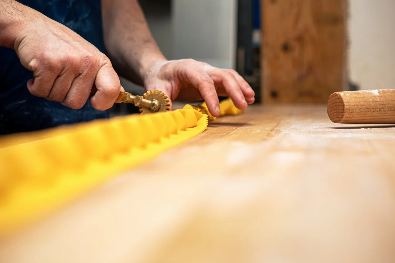 Person rolling out yellow pasta dough with a pastry wheel on a wooden countertop.