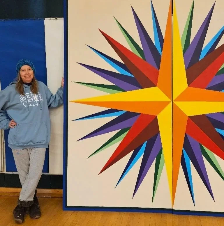 Woman in gray hoodie and beanie standing beside a large colorful geometric star mural painted on a wall.