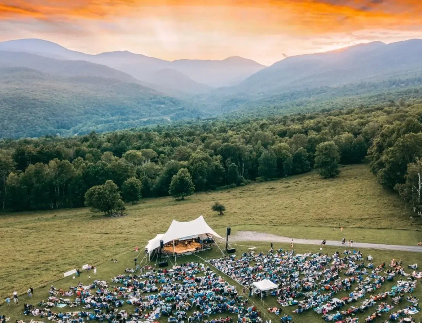 Outdoor concert with a large white tent stage, surrounded by numerous seated audience, set in a grassy field with mountains and trees in the background during sunset.