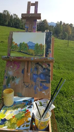 An outdoor painting setup with an easel holding a landscape painting of mountains, trees, and a blue sky, with painting supplies including a palette, brushes, and a cup of water on a table in front of a grassy field and trees in the background.
