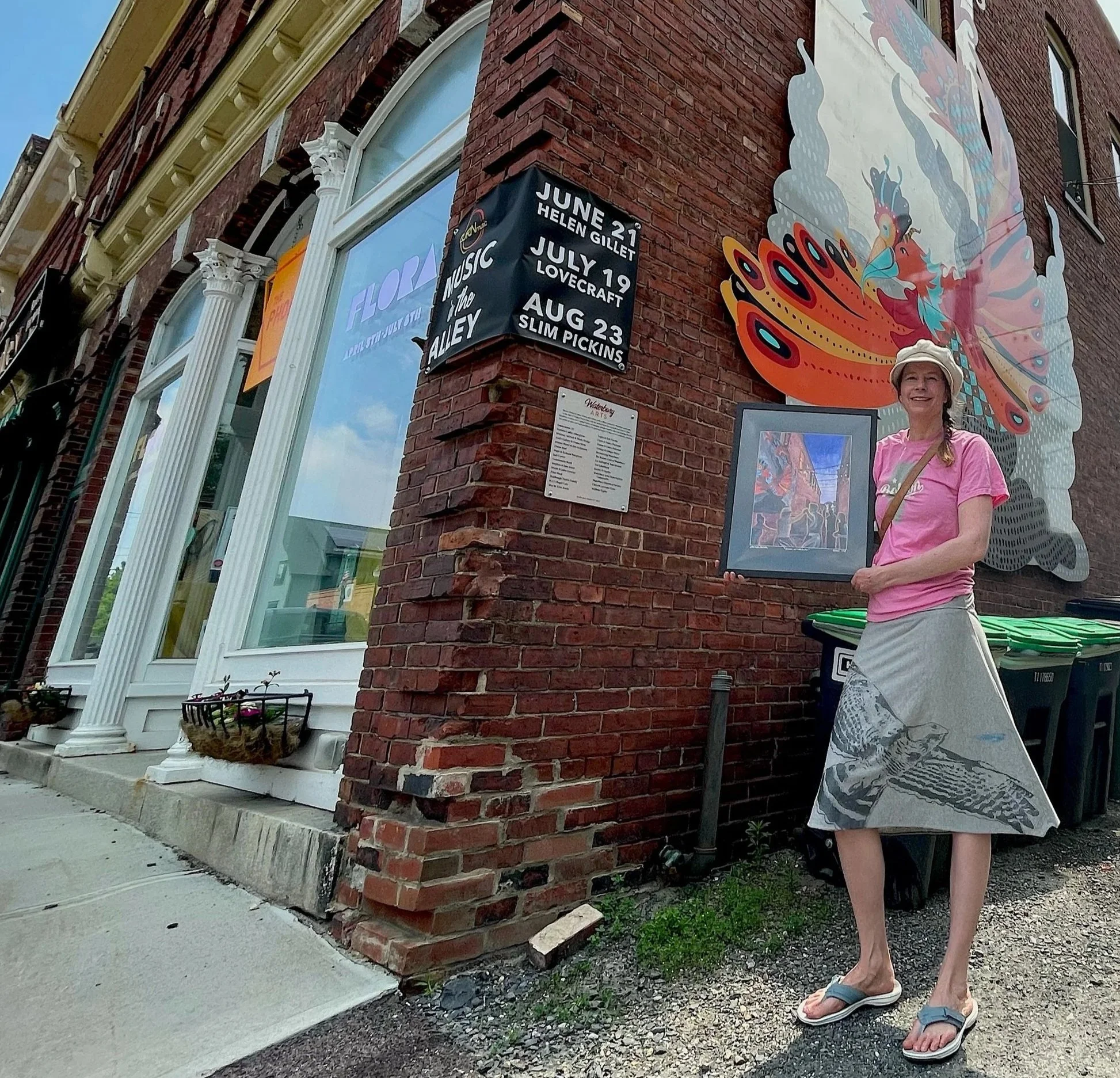 A woman in a pink t-shirt, white hat, patterned shorts, and flip-flops standing outside a brick building next to a corner with a decorated mural featuring a colorful butterfly. There is a black sign with white text listing upcoming events with dates in June, July, and August. The sidewalk has a flower basket and a small patch of grass beneath the woman.