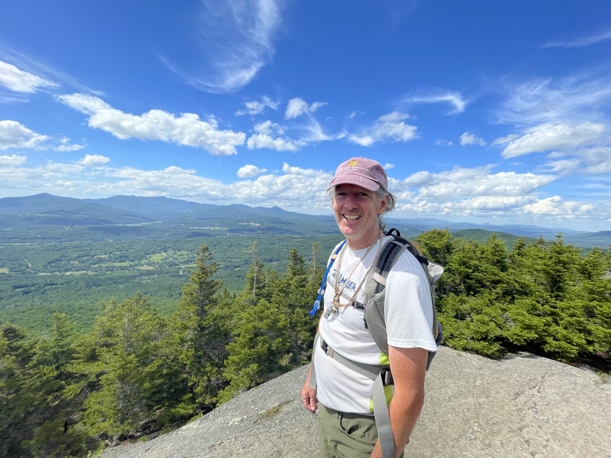 A man wearing a pink cap, white t-shirt, green shorts, and a hiking backpack stands outdoors on a hilltop amidst greenery, with a scenic view of mountains and a partly cloudy blue sky in the background.