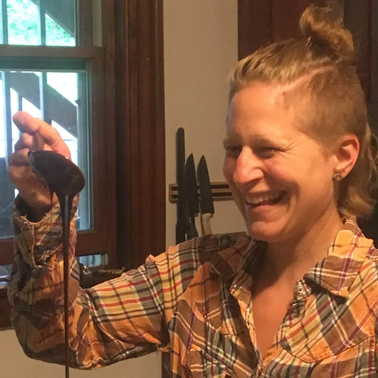 A woman smiling while holding up a small dark-colored animal, possibly a mole, in her hand in a kitchen with knives on a rack and a window in the background.