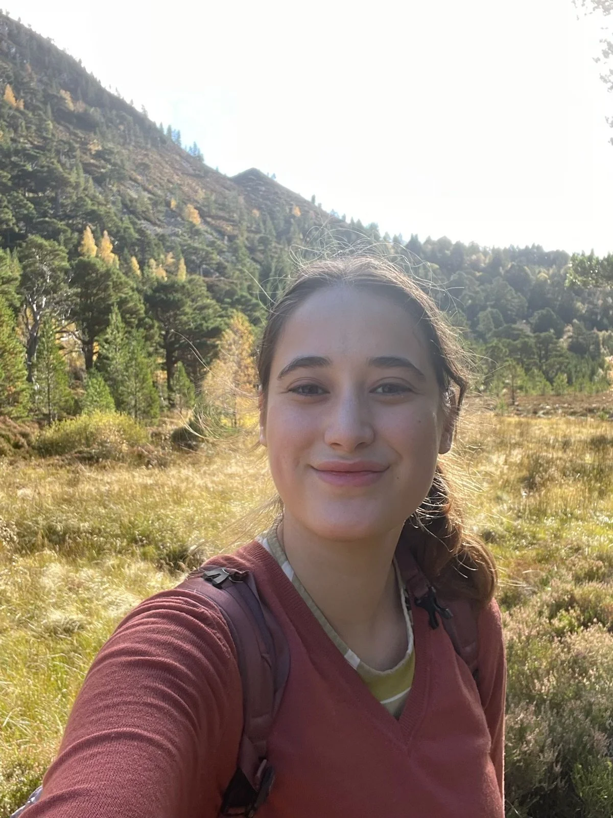 A young woman with brown hair in a ponytail smiling for a selfie outdoors in a grassy field with trees and mountains in the background on a sunny day.