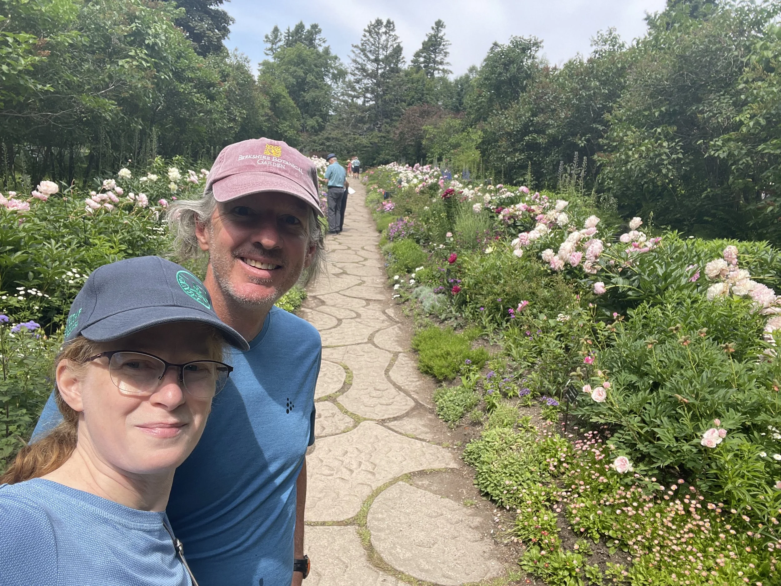 A group of three people, a man, a woman, and a child, taking a selfie in a garden filled with colorful flowers, with trees and sky in the background.