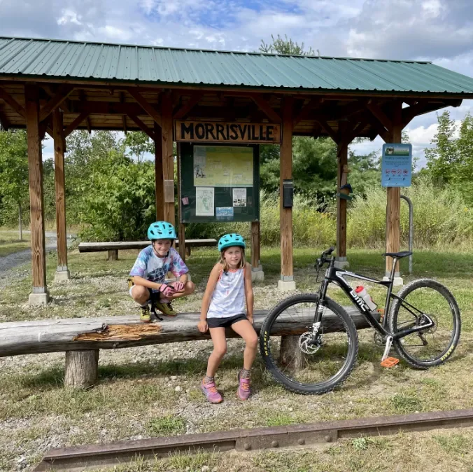 Two children wearing blue helmets sit on a wooden bench in front of a sign that reads 'Morrisville' at a park, with a bicycle leaning against the bench and nature in the background.