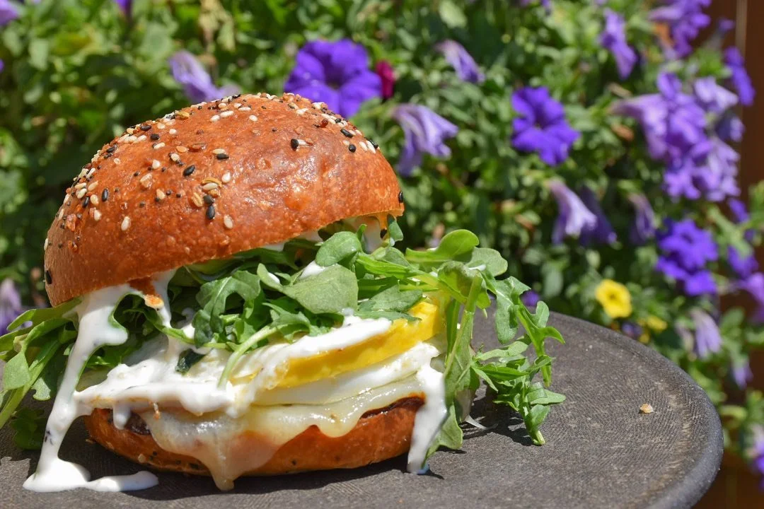 Sandwich with eggs, greens, and a multigrain bun on a black plate, with purple flowers in the background.