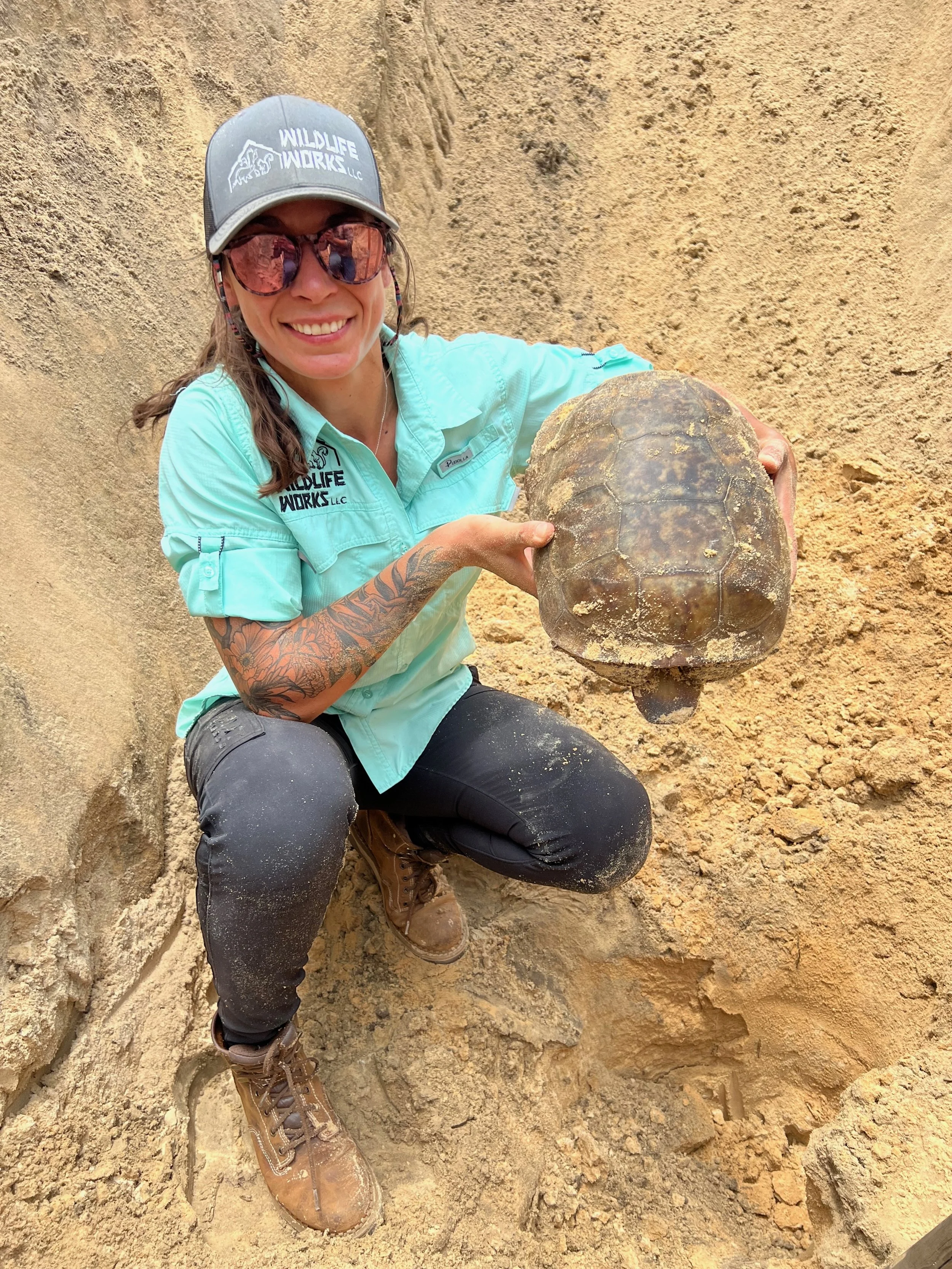 Woman crouching in a sandy area, holding a turtle shell, wearing sunglasses, a cap, and outdoor work clothes.
