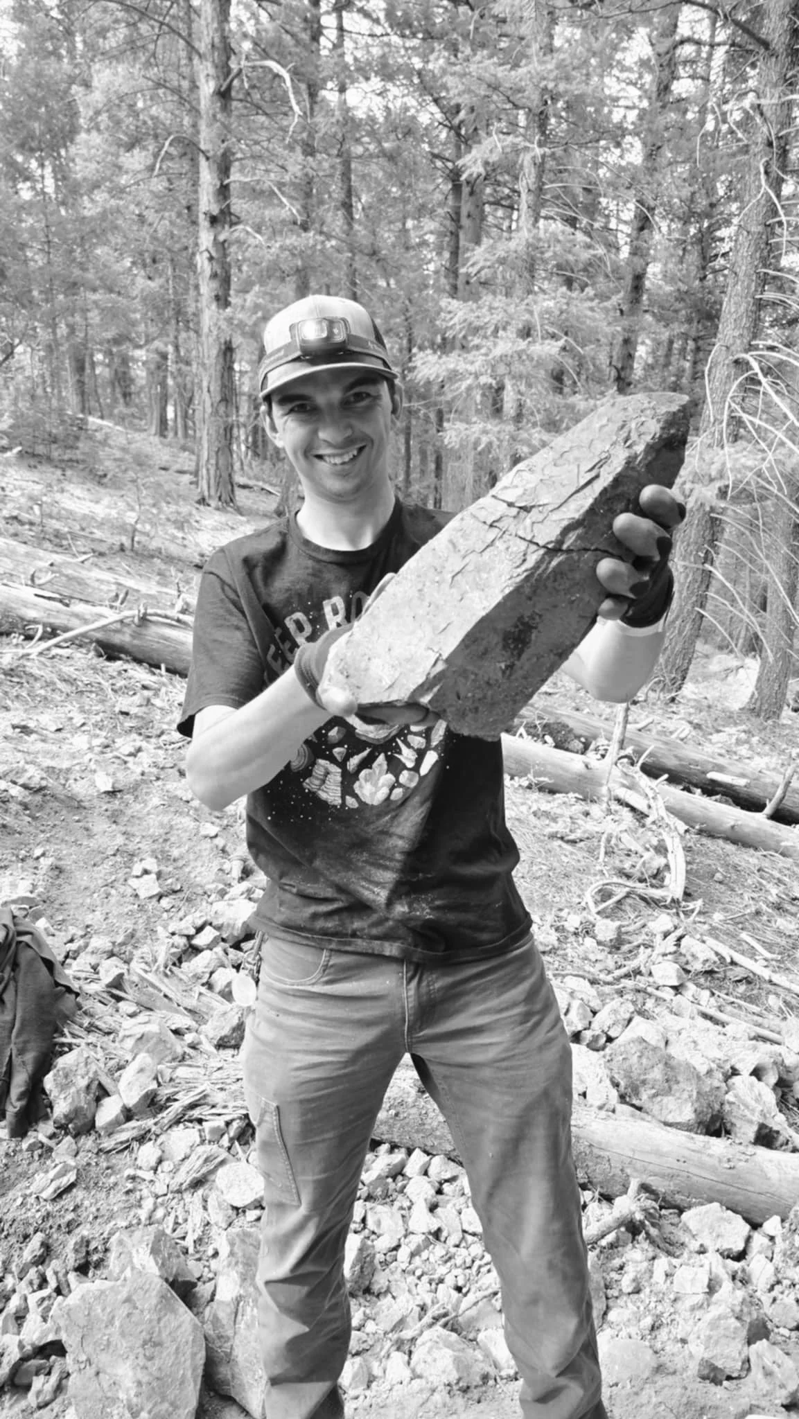 A man in a forest holding a large rock and smiling at the camera.