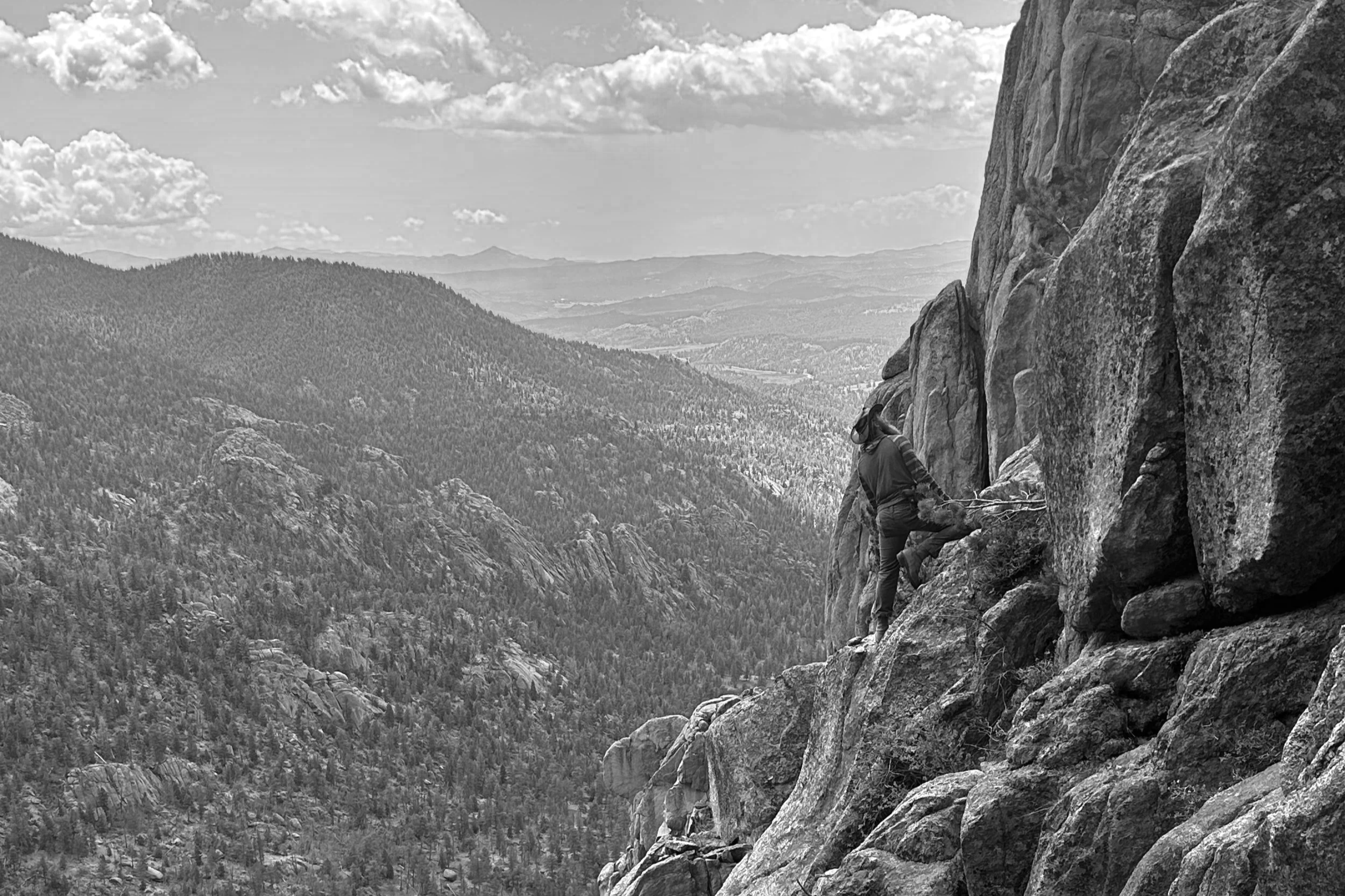 A person rock climbing on a steep cliff in a mountainous landscape with rolling hills and clouds in the sky.