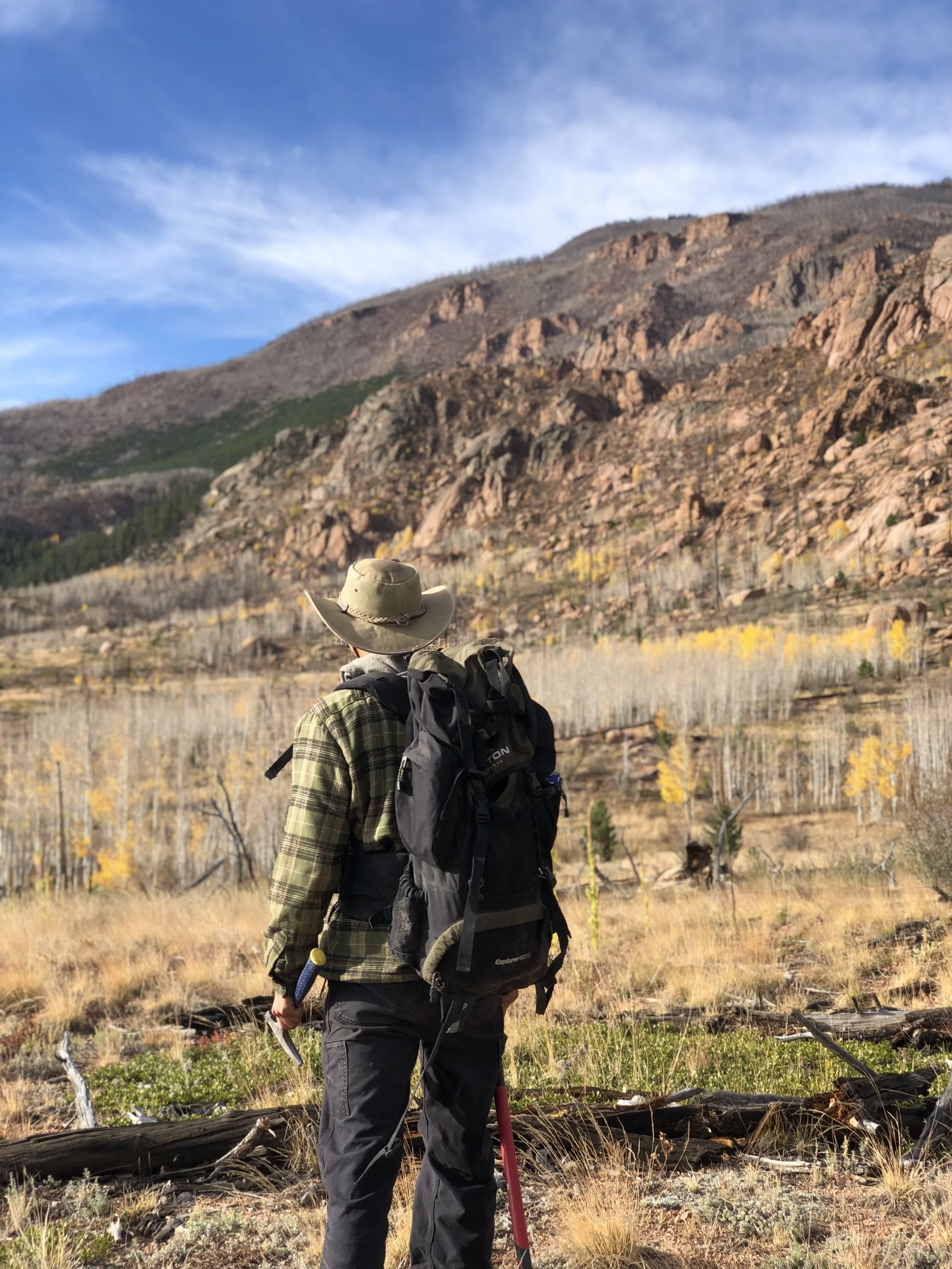 Hiker standing in a mountainous landscape with a backpack, wearing a wide-brimmed hat and plaid shirt, observing the scenery of ridges, rocky hills, and autumn trees under a partly cloudy sky.