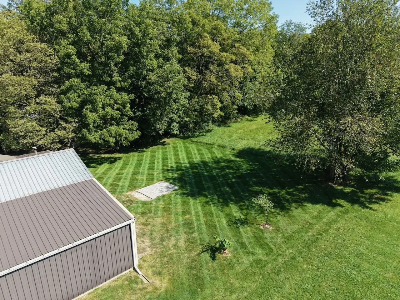 Aerial view of a backyard with a freshly mowed lawn, a metal building with a gray roof, trees, and a small garden area.