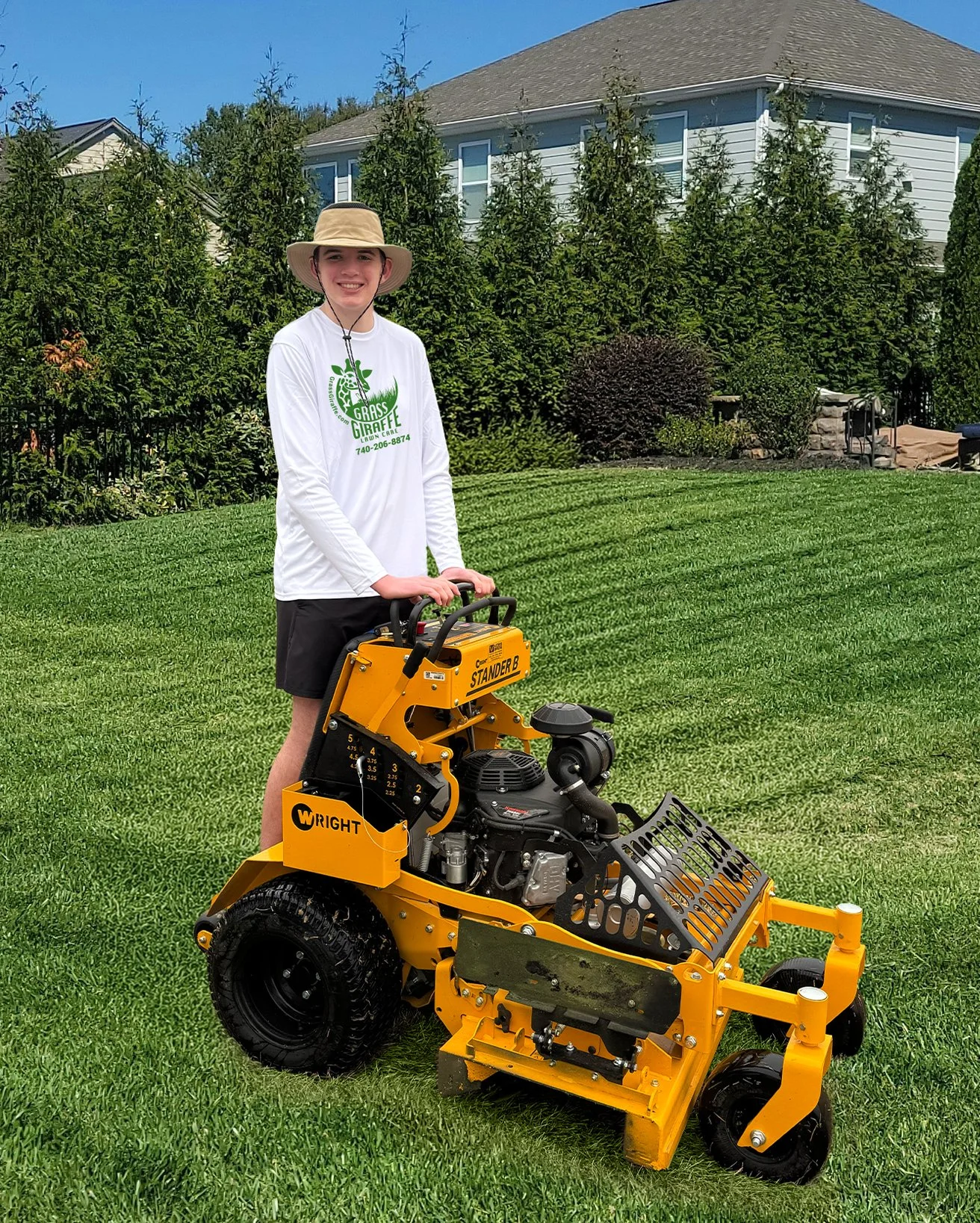 A young person standing on a yellow riding lawn mower in a backyard with green grass, tall trees, and a house in the background.