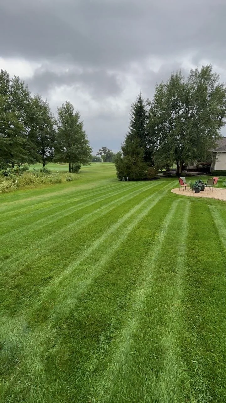 A well-maintained green lawn with mowing lines, trees, a patio with chairs, and a cloudy sky.