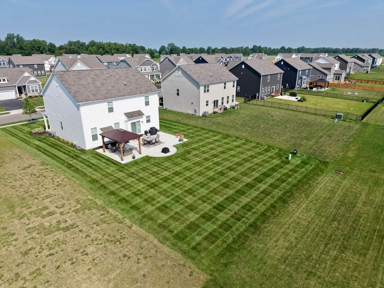 Aerial view of a suburban neighborhood showing several houses and their backyards, with one backyard featuring freshly mowed grass with a checkered pattern and outdoor furniture under a patio cover.
