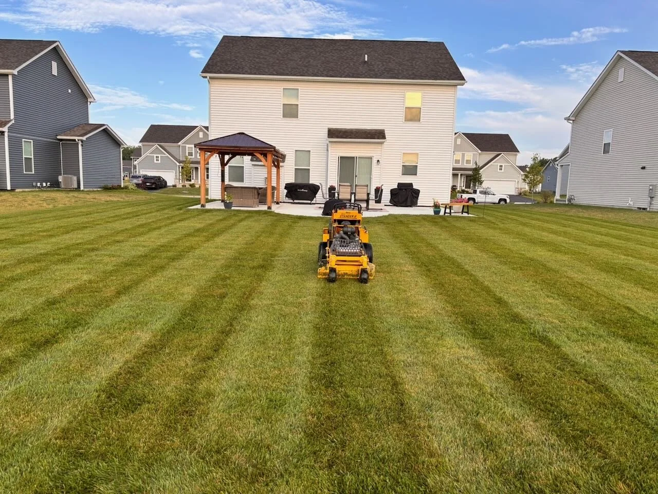 A backyard with freshly mowed lawn, a riding mower in the center, and a white house with outdoor furniture and a gazebo in the background.