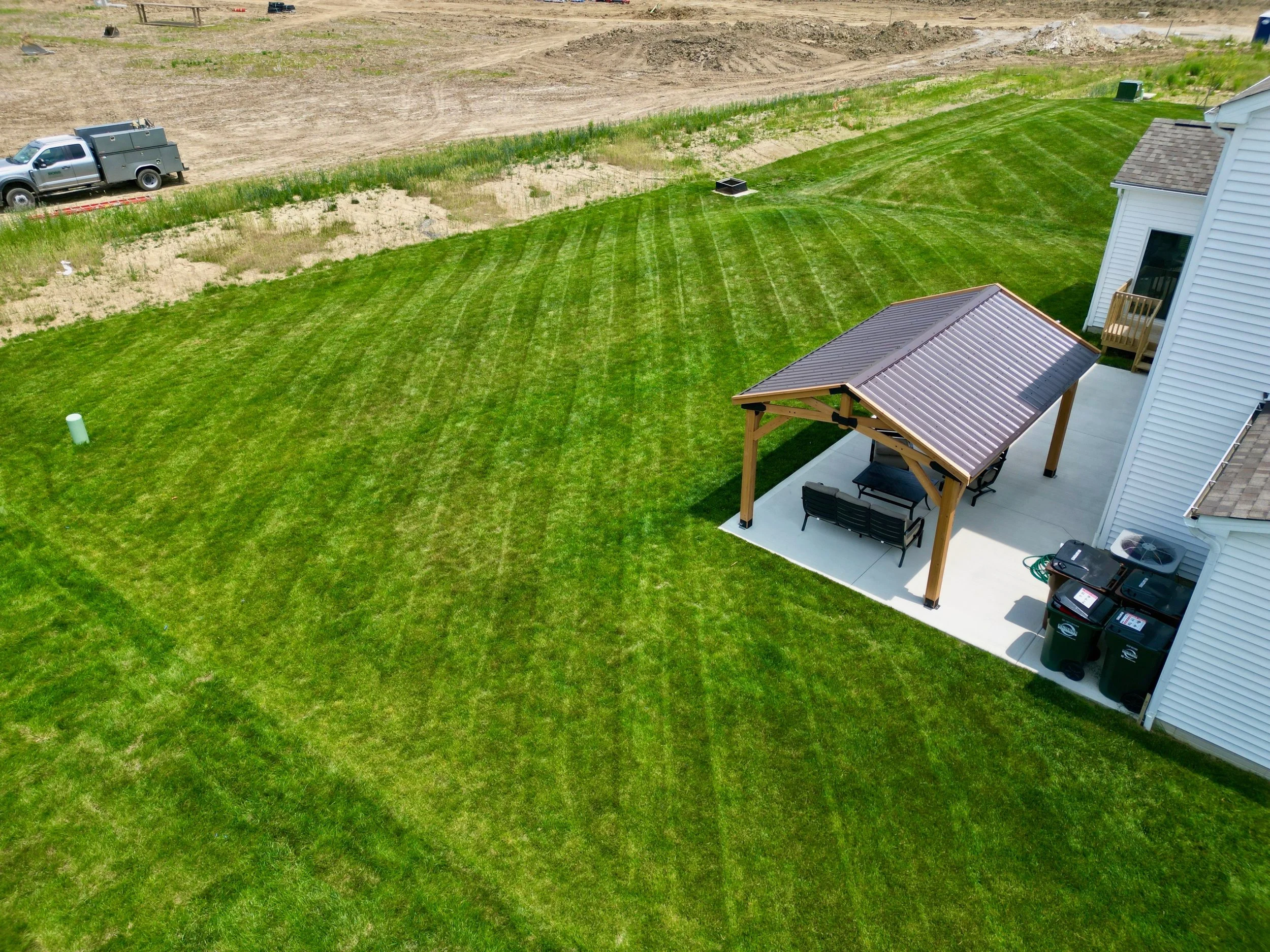 A backyard with a well-maintained green lawn, a white house with a wooden patio, a covered sitting area with two benches, and trash bins. There is also a truck with a utility utility bed parked near an area with dirt and grass.