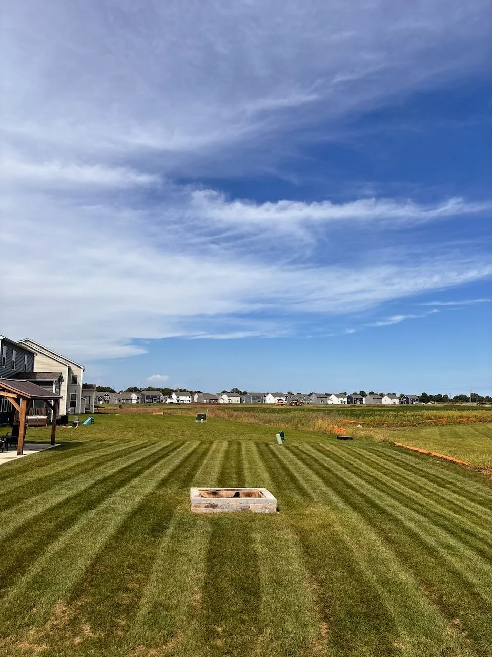 A large backyard with freshly mowed grass and visible striped mowing patterns, with a small fire pit made of bricks in the foreground, houses and a blue sky with clouds in the background.