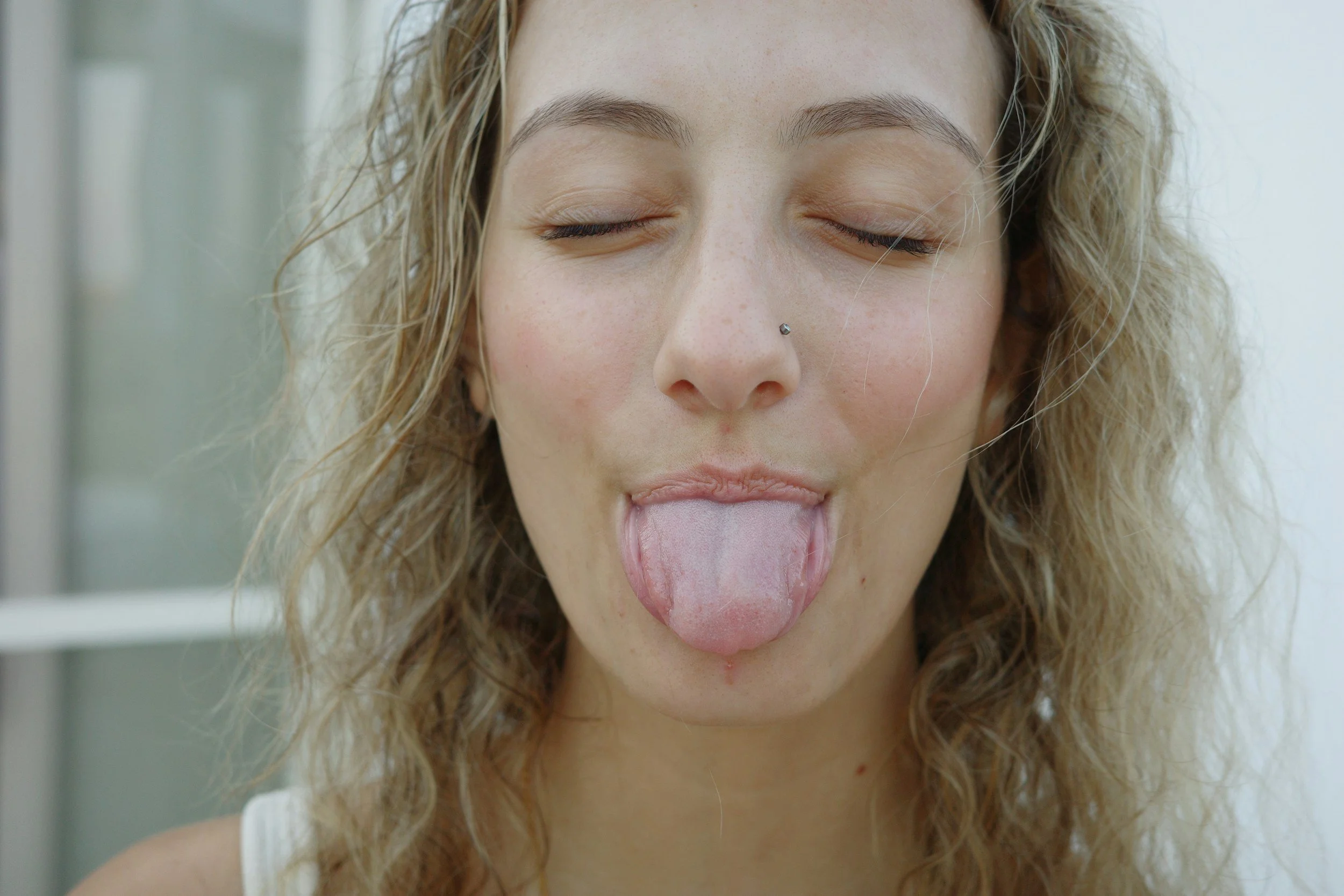 A woman with curly blonde hair and a nose piercing sticking out her tongue with her eyes closed.