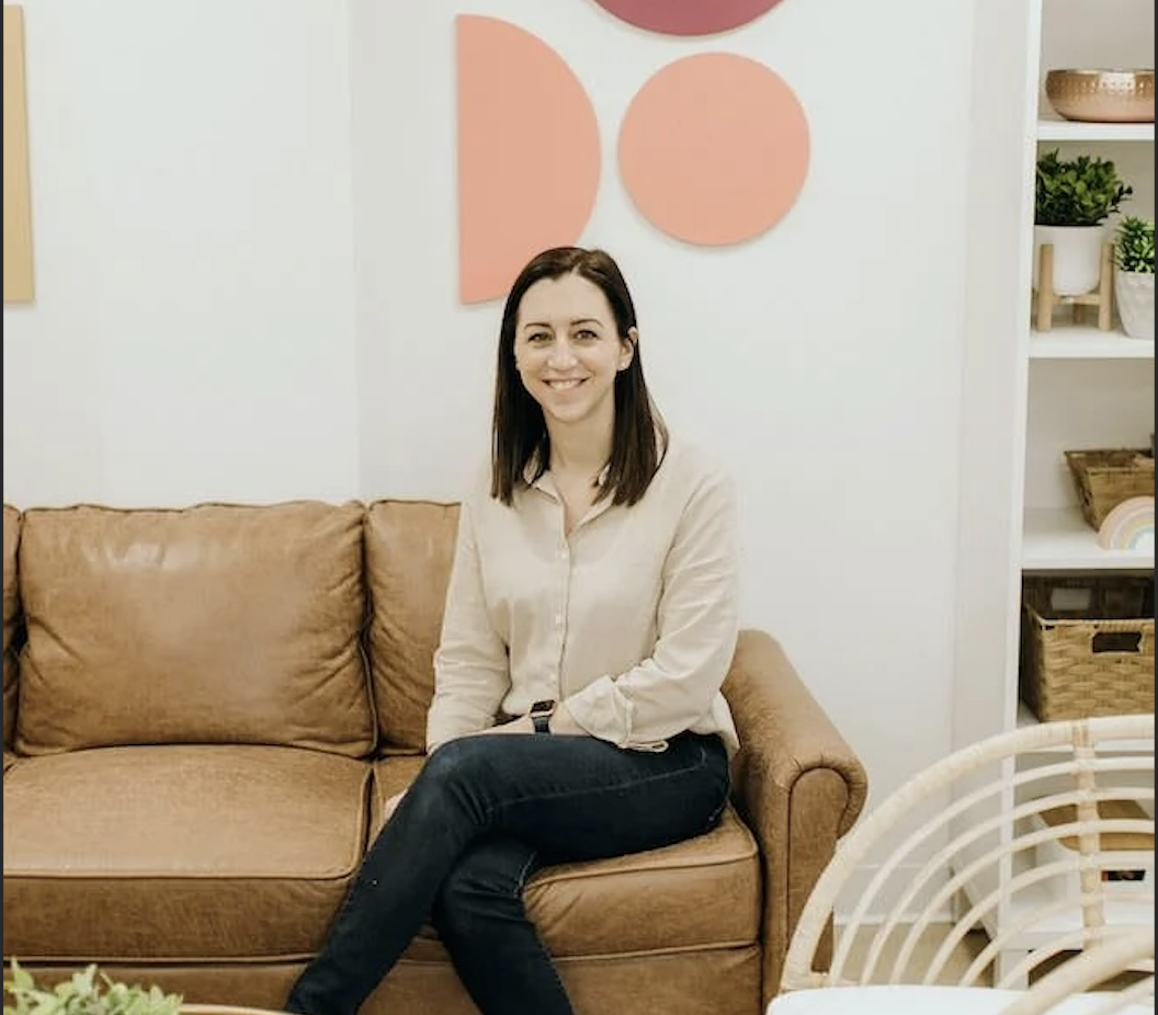 A woman sitting on a tan sofa in a room with modern decor, smiling at the camera, with pink geometric wall art and plants on shelves behind her.