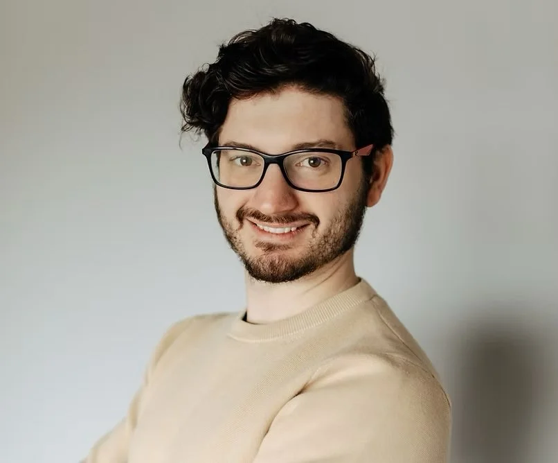A young man with curly dark hair, glasses, and a beard smiling at the camera, wearing a beige shirt against a plain light gray background.