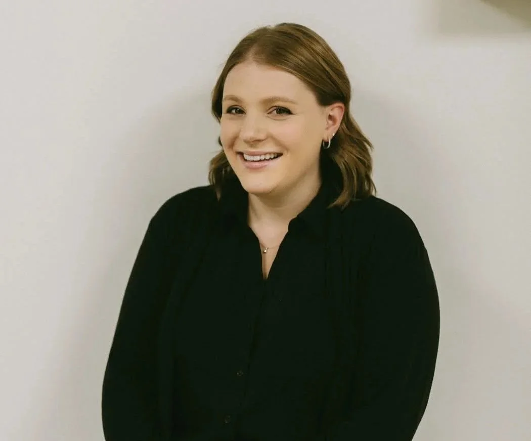 A woman with shoulder-length brown hair wearing a black top and small earrings, smiling and standing against a plain light-colored wall.