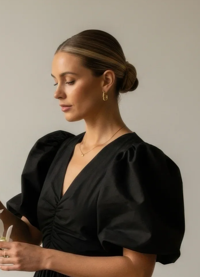 A woman with styled hair, wearing gold earrings and a delicate necklace, dressed in a black dress with puffy sleeves, holding a paper bag.